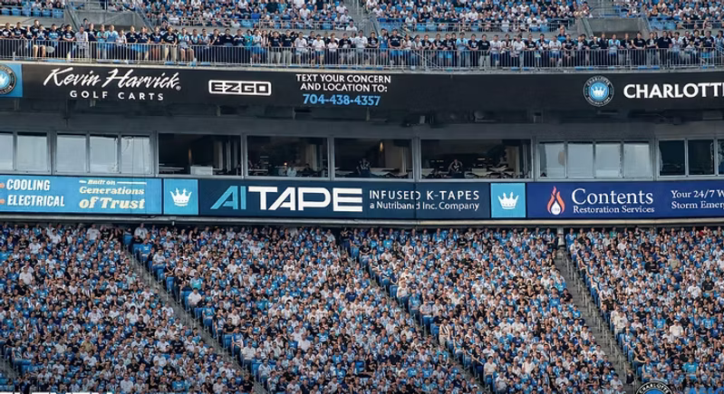 AI Tape stadium advertising banner displayed on the stands during a Charlotte Football Club match with crowds of fans.