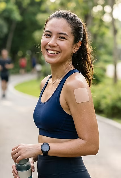 A smiling athletic woman jogging outdoors in a park, wearing a beige Active Intelligence All-Day Energy Patch on her left shoulder.