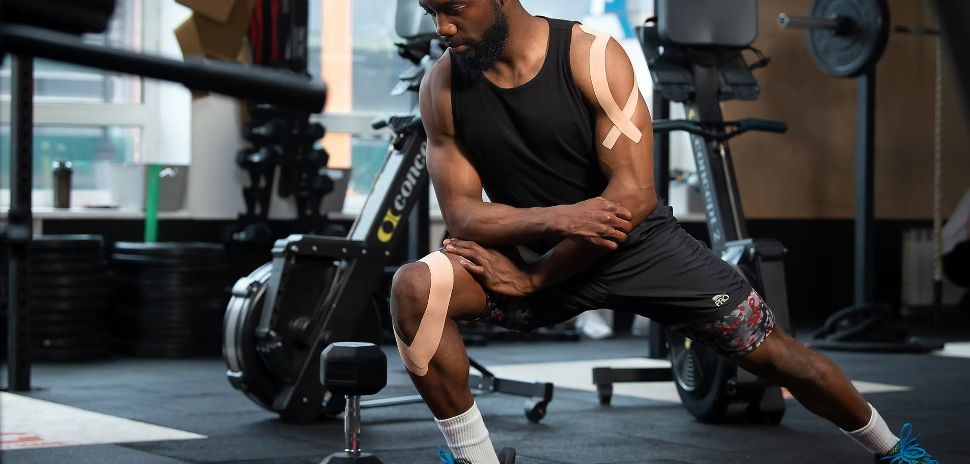 An athletic man stretching inside a fitness gym, showcasing applied beige Active Intelligence kinesiology tapes on his shoulder and knee.