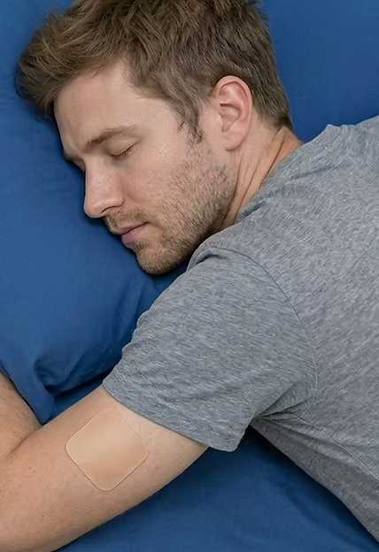 A young man sleeping peacefully on a blue pillow, wearing a beige Active Intelligence Sleep & Recovery Patch on his upper arm.