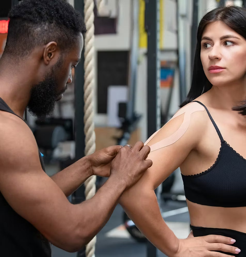 A male trainer applying beige Active Intelligence kinesiology tape to a woman's shoulder inside a gym setting.